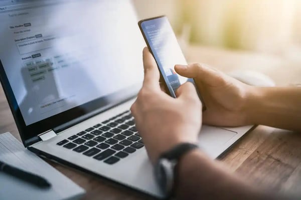 Personne utilisant un smartphone devant un ordinateur portable, illustrant la navigation et la gestion du consentement sur plusieurs appareils (multi-écrans).
