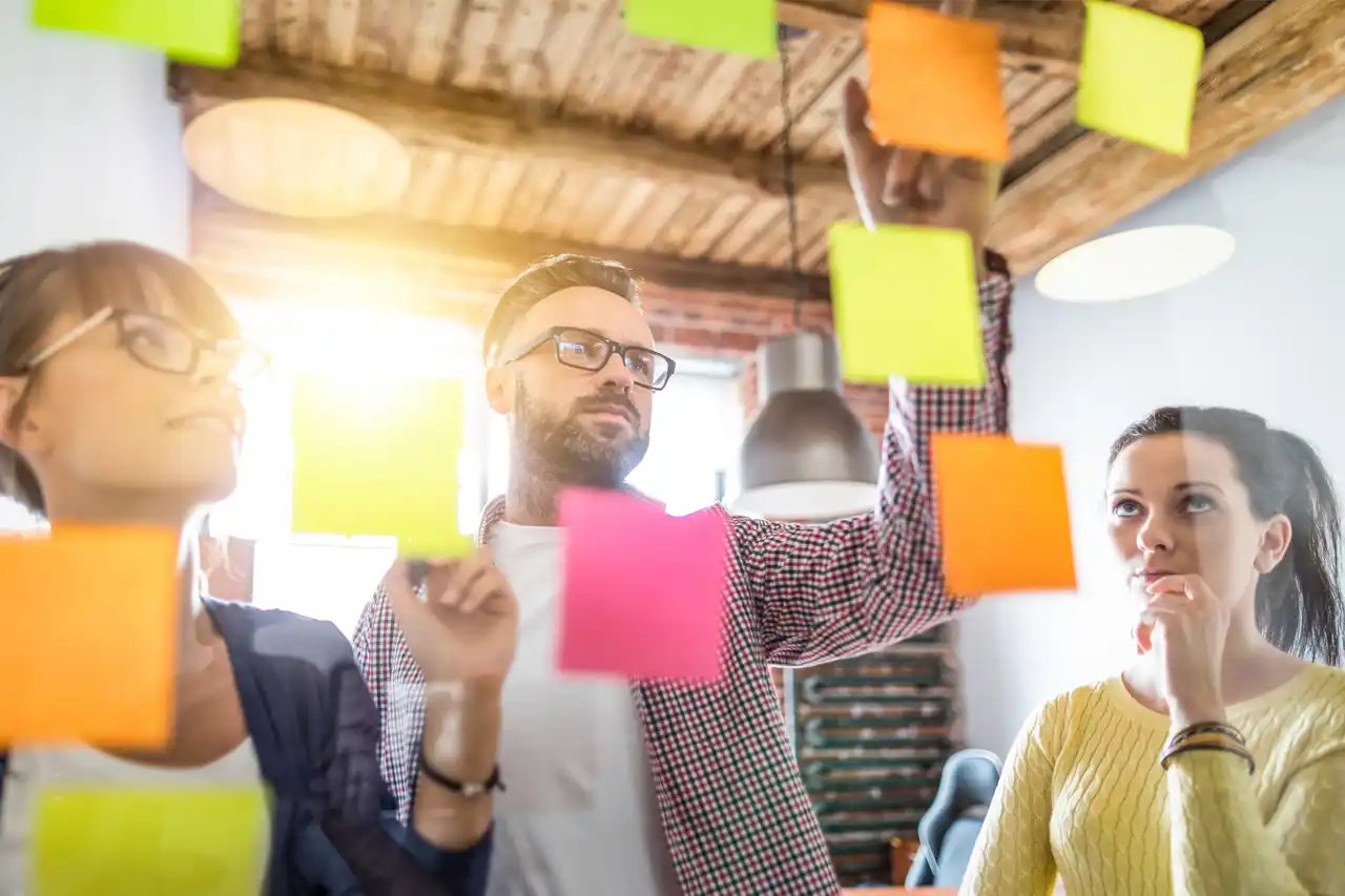 Un groupe de travail en plein atelier de design thinking. Deux femmes et un homme positionnent, échangent concernant des post-its accrochés au mur.